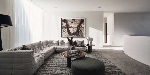 A bright, spacious contemporary living room with a large low-profile modular sectional sofa arranged around a dark stone or glass coffee table and round ottoman. Floor-to-ceiling sheer curtains line the left side, while a textured grey carpet, pale walls, recessed ceiling lights, abstract artwork, sculptural decor, and a curved white architectural partition create a refined minimalist setting. A hallway or adjacent space is visible beyond the open doorway.
