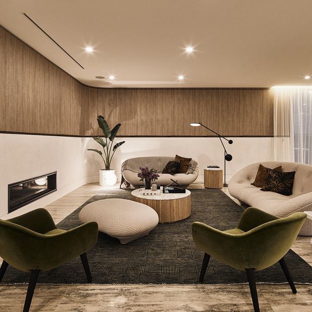 A warm, refined living room with curved built-in wood wall panelling, a low linear fireplace, sculptural upholstered sofas and armchairs, a round wood coffee table, and layered rugs. Recessed ceiling lights and sheer curtains create a softly lit, intimate atmosphere.