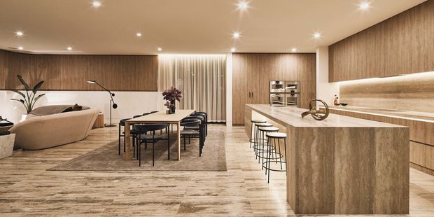 A spacious open-plan contemporary interior combining kitchen, dining and living areas. The kitchen features a large stone island with integrated seating, timber-look cabinetry, a built-in appliance wall and a softly lit backsplash. A dining table and chairs sit centrally on a rug, while a curved sofa and lounge area are visible beyond. The room has warm layered lighting, continuous timber-toned finishes and light stone-look flooring.