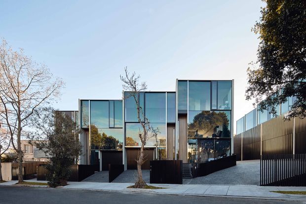 Street-facing exterior view of a contemporary multi-level residence with a strongly rectilinear composition, extensive floor-to-ceiling glazing, dark vertical cladding, and projecting box-like volumes. The home sits behind a paved forecourt with dark fencing and a sloping driveway, with mature trees and neighboring houses visible around it.