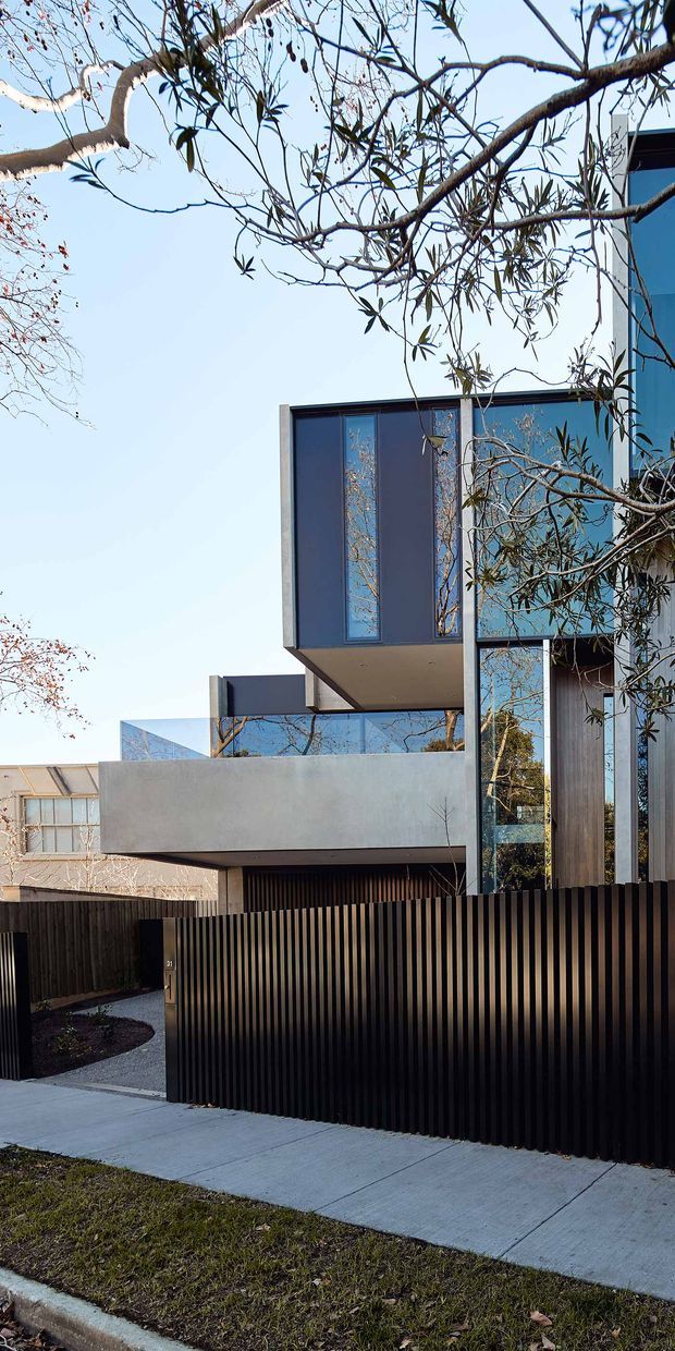 Exterior street-level view of a contemporary multi-storey residence with projecting box forms, extensive dark-framed glazing, smooth pale rendered walls, and a tall black vertical-slat front fence. Mature tree branches partially frame the facade, while a concrete footpath and landscaped entry sit in the foreground.