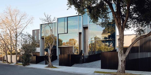 Street-facing view of a contemporary multi-storey residence with a strongly rectilinear composition, extensive floor-to-ceiling glazing, dark vertical metal fencing, and a concrete-paved driveway. Mature street trees partially screen the facade, while the glazing reflects the surrounding neighbourhood and sky.
