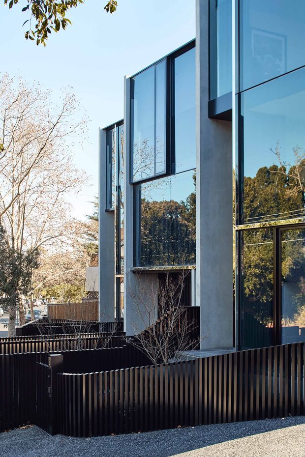 Exterior view of a contemporary multi-storey residence with a strong vertical composition, tall dark-framed glazing, pale concrete or rendered structural walls, and reflective floor-to-ceiling windows. A dark vertical-slat front fence and landscaped planting define the street edge, while trees and neighbouring residential context are reflected in the glazing.