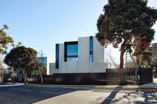 Street-facing view of a contemporary multi-level residence with a crisp geometric facade, pale rendered or concrete-like wall volumes, dark vertical metal fencing, and large blue-tinted glazed openings. Mature trees frame the property, with a paved footpath and quiet suburban road in the foreground.