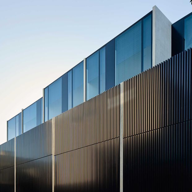 A close-up exterior architectural view showing a contemporary facade composed of tall blue-tinted glazed panels, pale concrete vertical structural fins, and a lower screen of closely spaced dark vertical slats. The linear composition emphasizes the building envelope and strong geometric repetition against a clear sky.