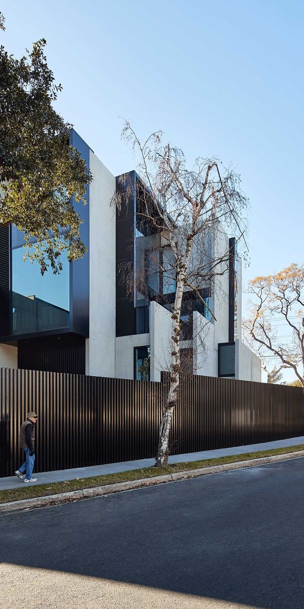 Street-level exterior photograph of a contemporary multi-storey residence with strong geometric massing, white rendered walls, dark charcoal cladding and extensive dark-framed glazing. A tall ribbed black privacy fence fronts the property, with a narrow footpath, street and mature trees in the foreground.