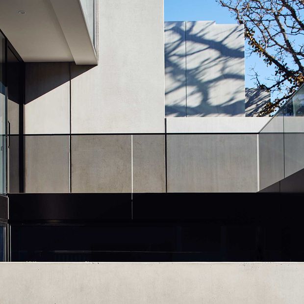 A minimalist modern architectural exterior detail with strong horizontal and vertical geometric lines. The facade combines pale smooth concrete or rendered wall panels, dark charcoal cladding, extensive dark glazing, and a transparent glass balustrade. Sharp sunlight creates tree-shadow patterns across the upper wall, while a projecting roof or overhang adds depth and contrast.