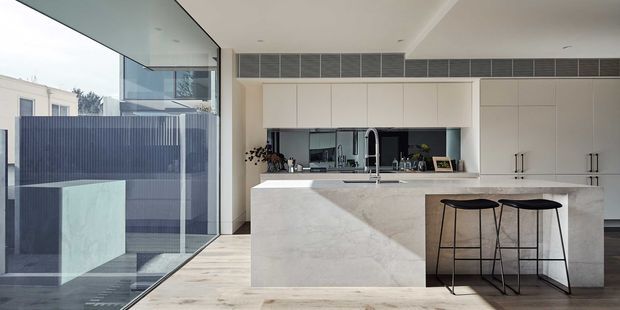 Contemporary kitchen with a large pale stone island and integrated seating, full-height white cabinetry, dark reflective splashback, and a timber-look floor. Floor-to-ceiling glazing opens the kitchen visually toward an exterior courtyard or terrace, while a linear ceiling ventilation grille adds a functional architectural detail.