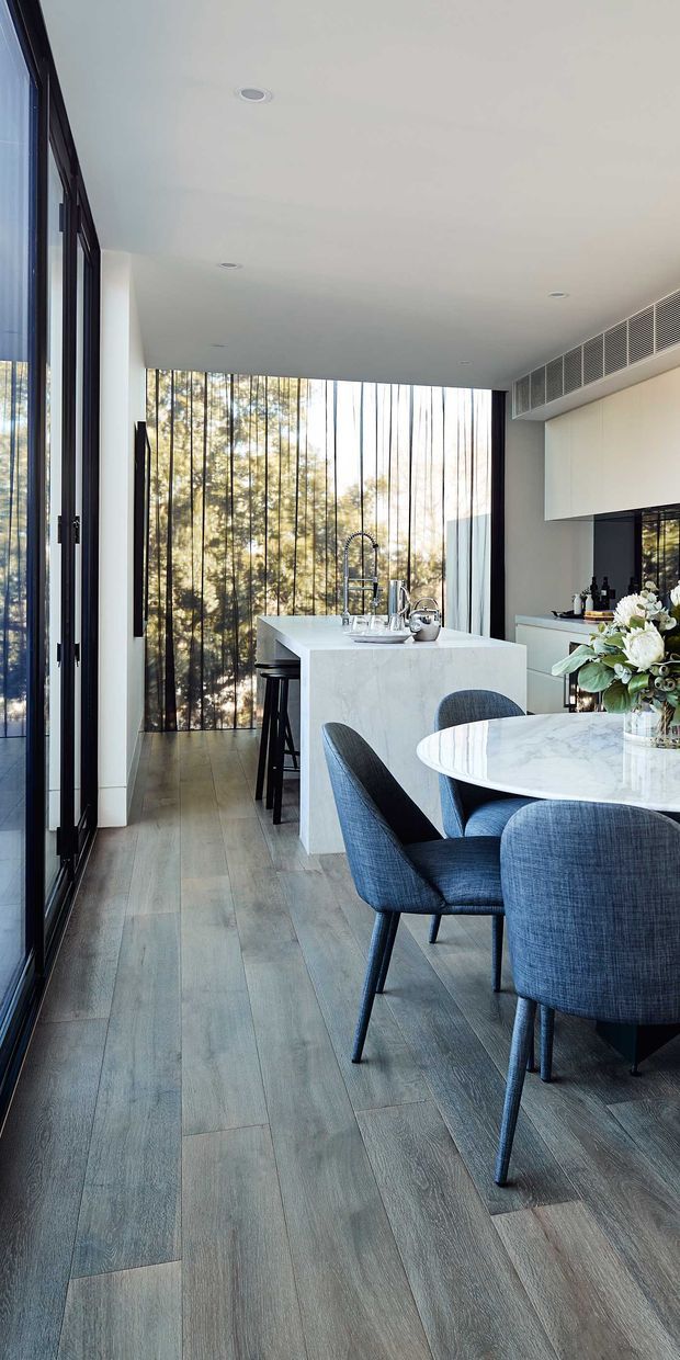A bright, contemporary open-plan kitchen and dining area with full-height dark-framed glazing along one side and a wall of vertical sheer curtains overlooking trees. The room features a white stone kitchen island with integrated seating, streamlined white cabinetry, a pale timber-look floor, and a round white stone dining table surrounded by blue upholstered chairs. Recessed ceiling lighting and a linear ceiling air-conditioning vent maintain a clean, refined aesthetic.