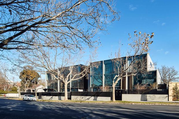 Street-facing view of a large contemporary multi-level residence with a strongly glazed facade, dark vertical cladding or framing, light concrete boundary walls, and a flat roofline. Mature deciduous street trees partially screen the building, with a paved road and driveway frontage in the foreground under clear blue sky.