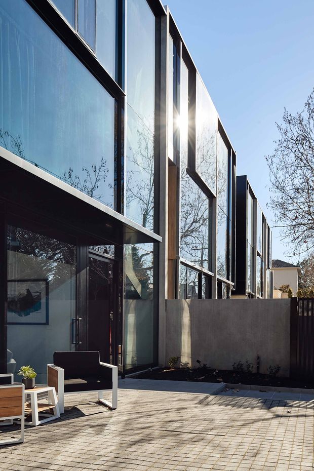 Exterior view of a contemporary multi-storey building with a dramatic glazed facade framed by dark metal and vertical timber elements. The facade opens onto a paved courtyard or terrace with outdoor seating, low rendered boundary walls, and mature deciduous trees reflected in the extensive glazing. Strong sunlight creates a crisp, high-contrast architectural photograph.