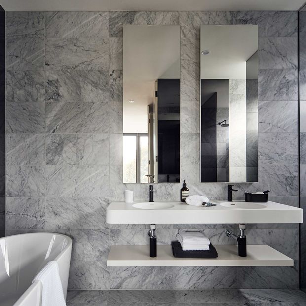 Contemporary bathroom with floor-to-ceiling grey marble-look wall tiling, a floating white double vanity with black tapware, twin rectangular mirrors, neatly folded towels, and a freestanding white bathtub partially visible in the foreground. The space has a refined monochromatic palette and clean-lined fixtures.