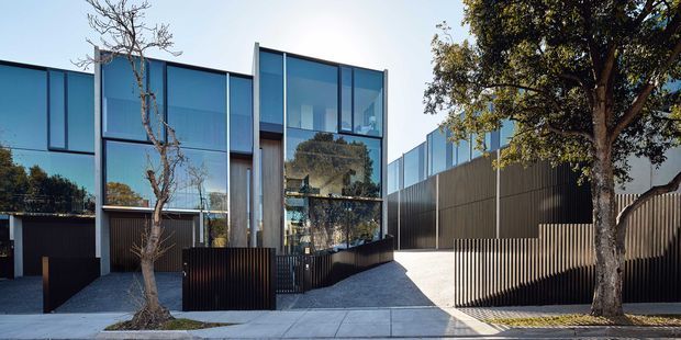 Street-facing view of a contemporary multi-level residence with a strongly glazed facade, dark vertical metal or timber-like screening, projecting box forms, and a recessed entry. The property is set along a suburban sidewalk with landscaped trees and a gravel-paved forecourt. Reflective floor-to-ceiling windows reveal only limited interior detail.