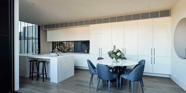A bright, refined open-plan kitchen and dining area. The space features a large pale stone island with integrated sink and seating, extensive full-height white built-in cabinetry, dark timber-look flooring, floor-to-ceiling glazing, and a round dining table surrounded by blue upholstered chairs. A linear ceiling ventilation grille and minimalist recessed lighting contribute to the contemporary design.