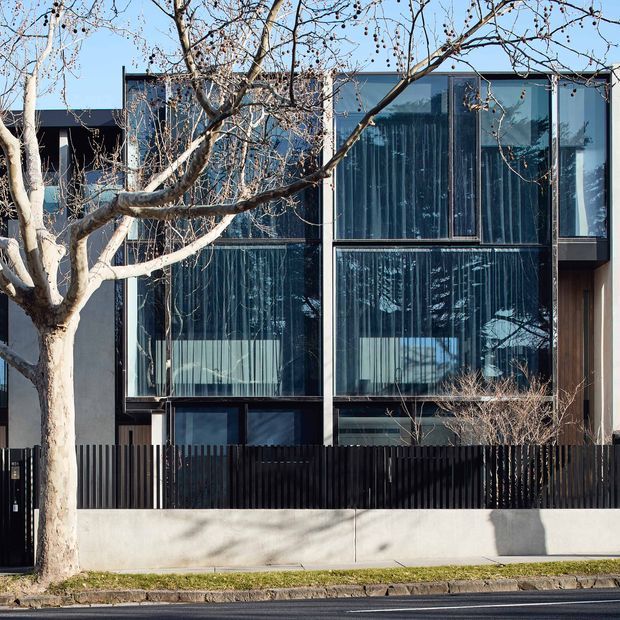 Street-facing view of a contemporary multi-storey townhouse or residential building. The facade combines extensive floor-to-ceiling dark-framed glazing, dark vertical metal fencing, pale rendered walls, and a simple flat roofline. A mature deciduous street tree partially obscures the facade.