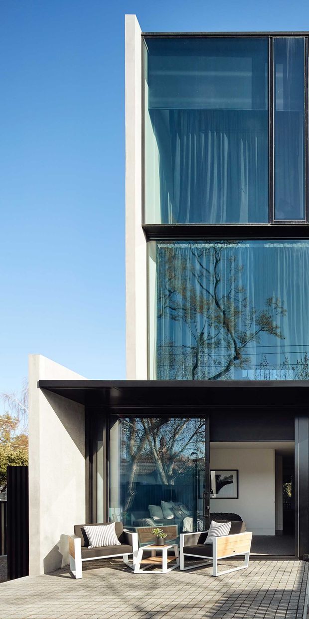 A close exterior view of a multi-storey contemporary home with a strongly geometric facade. Large floor-to-ceiling glazed openings with dark metal frames dominate the upper level, while white rendered wall planes and black horizontal roof or canopy elements create sharp contrast. A paved terrace with outdoor lounge chairs is visible at ground level, and trees are reflected in the glazing.