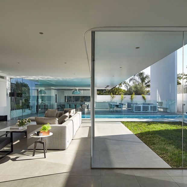 A bright, expansive contemporary living area with large frameless glass walls opening directly to a covered terrace and swimming pool. The room contains a large pale sectional sofa, coffee table, television wall, and sculptural pendant lighting. The exterior view includes a tiled terrace, lawn, pool, outdoor dining area, white architectural columns, and tropical planting. The space has a strong indoor-outdoor connection and abundant natural light.