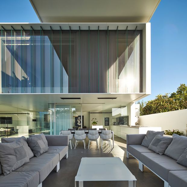 Bright contemporary two-storey residential living and dining space opening to an outdoor courtyard. The image is viewed from the living area toward a white kitchen and dining zone, with extensive floor-to-ceiling glazing, a dramatic projecting upper volume, and vertical screened glazing creating strong linear shadows. Two large grey sofas and a white coffee table occupy the foreground, while a dining table and kitchen island sit beyond.