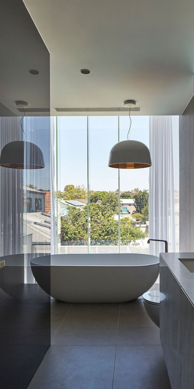 A refined contemporary bathroom with a freestanding oval bathtub positioned in front of full-height glazed windows. The room has dark large-format tiled flooring, a dark stone or tile wall on the right, a minimalist vanity, a partially visible toilet, recessed ceiling lighting, and two large grey pendant lights. Sheer curtains filter daylight while providing views toward a leafy suburban setting.