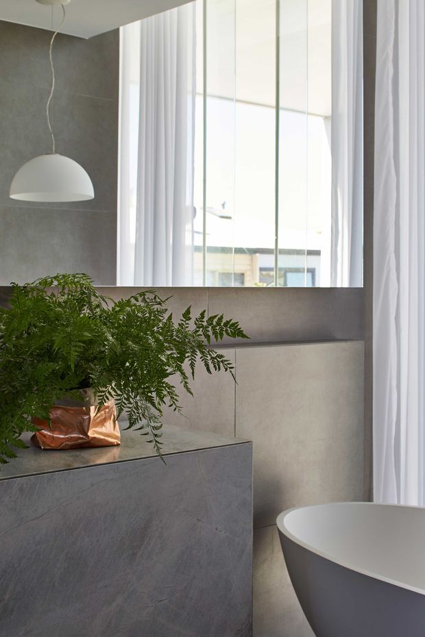 A refined contemporary bathroom featuring a freestanding white bathtub, large frameless mirror, floor-to-ceiling sheer curtains, grey stone-look wall and cabinetry surfaces, and a sculptural white pendant light. A leafy fern in a metallic planter adds an organic accent to the otherwise calm, minimalist space.