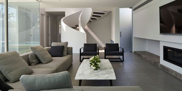 A bright, spacious contemporary living room with floor-to-ceiling glazing, a neutral sectional sofa, two black-framed lounge chairs, a marble-look coffee table, and a sleek linear fireplace beneath a wall-mounted television. A sculptural curved staircase forms a strong architectural feature at the rear of the room, with dark tiled flooring and crisp white walls and ceilings.