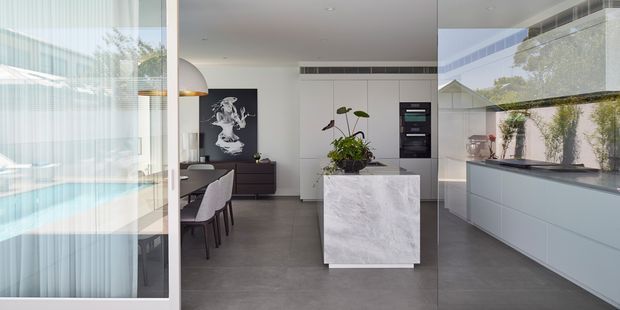 Contemporary open-plan kitchen and dining area viewed through large glazed sliding doors. The space features a pale grey island with a white marble-look surface, integrated pale cabinetry, dark grey flooring, a dining table with upholstered chairs, and a black artwork wall. Extensive glazing creates reflections and views toward a bright outdoor pool area. A tall indoor planter adds greenery to the restrained, monochromatic interior.
