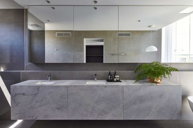 Contemporary minimalist bathroom vanity with a long floating grey stone benchtop and integrated cabinetry, double basins, large mirrored wall cabinets, dark grey tiled walls, recessed ceiling lights, and a freestanding white pendant. A plant adds a small natural accent, while a partial freestanding bathtub is visible at the right edge.