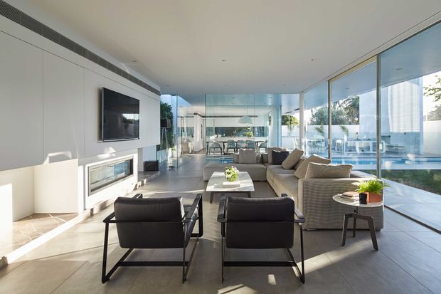 A bright, spacious contemporary living room with floor-to-ceiling glazing on two sides, opening views toward a pool and landscaped exterior. The room features a streamlined white built-in media wall with fireplace, pale tiled flooring, a large neutral sectional sofa, two dark leather lounge chairs, a low coffee table, and a dining area visible beyond. Strong daylight and reflective glazing create an airy, polished atmosphere.