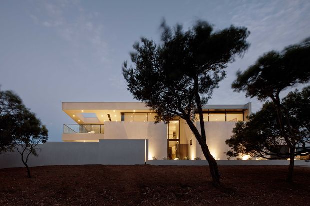 Twilight exterior view of a contemporary, flat-roofed multi-level residence set behind a low boundary wall. The home features clean white rendered volumes, extensive floor-to-ceiling glazing, illuminated recessed soffits, balconies, and warm interior lighting. Dark sculptural trees frame the facade and the landscaped foreground is subdued in low light.