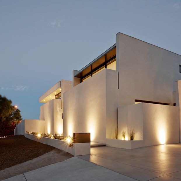 Twilight exterior view of a large minimalist modern residence with crisp geometric white rendered volumes, recessed openings, integrated warm architectural lighting, and a broad concrete driveway. The design includes a flat roof, projecting upper-level forms, dark-framed glazing, low boundary walls, and sparse architectural landscaping.