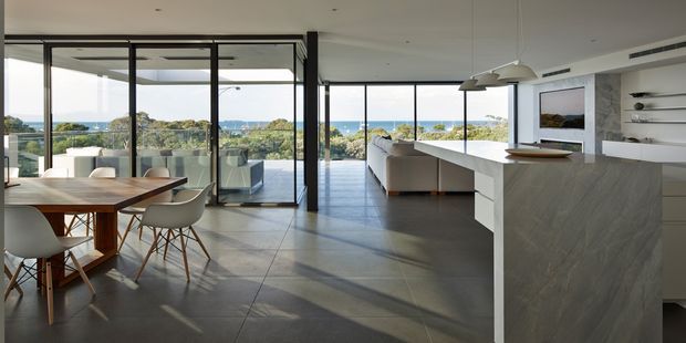A bright, open-plan contemporary kitchen, dining, and living area with expansive floor-to-ceiling glazing overlooking a coastal landscape. The kitchen features a large white island with a grey stone-look waterfall benchtop, integrated cabinetry, open shelving, and sculptural pendant lights. A timber dining table with modern upholstered chairs occupies the foreground, while a glazed outdoor or sunroom area extends to the left. Large-format grey floor tiles and black-framed glazing create a clean, refined interior.