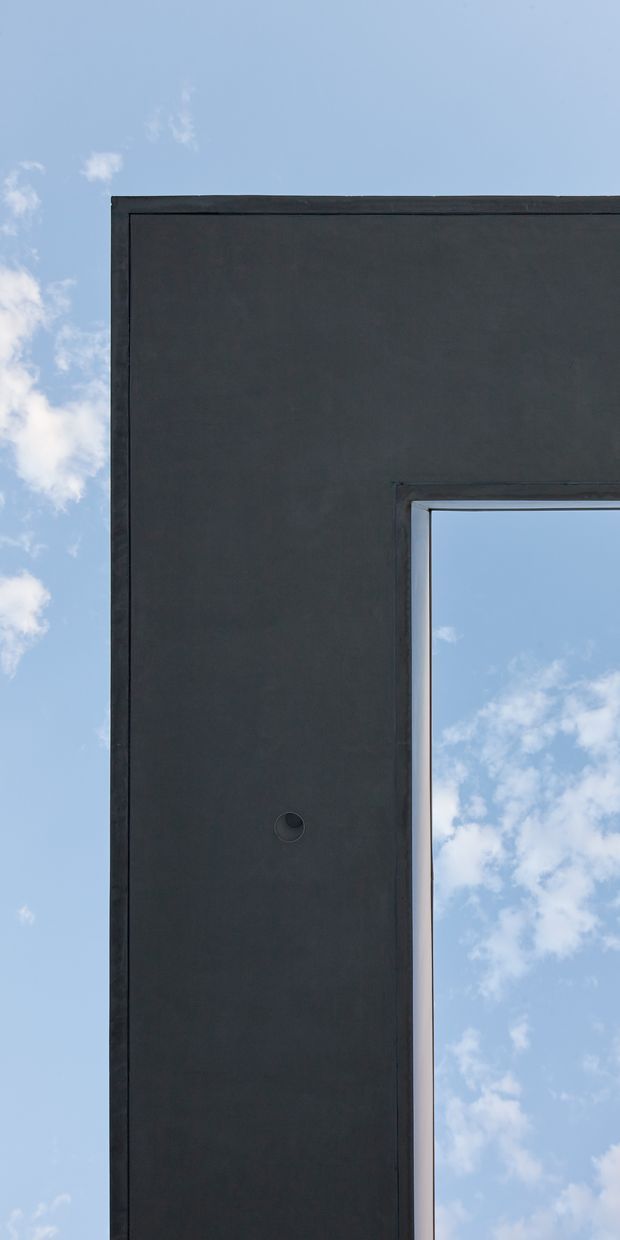 A close-up architectural exterior detail showing a dark charcoal, smooth wall or facade panel with a sharply recessed rectangular opening. The opening is edged with a slim dark frame and reveals blue sky and clouds beyond, creating a stark geometric composition with minimal detailing.