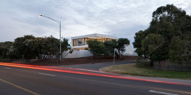 A dusk exterior view of a contemporary two-storey building set behind a landscaped verge on a suburban street. The building has a simple rectilinear form, a flat roof, extensive upper-level glazing, light-coloured rendered walls, and a recessed illuminated terrace or balcony. Long-exposure red vehicle light trails run along the road in the foreground, with mature trees and street lighting surrounding the property.