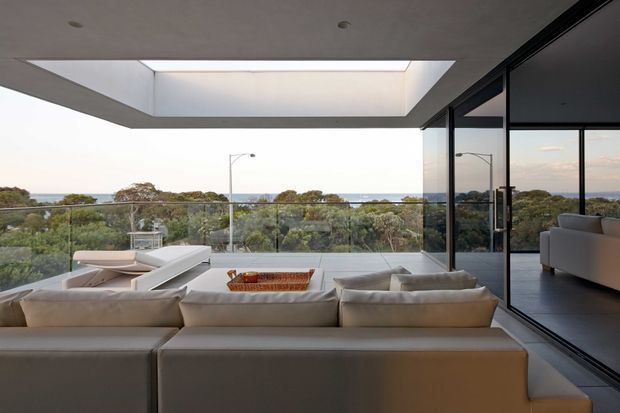 A contemporary elevated living area with expansive floor-to-ceiling glazing and a frameless glass balustrade overlooking a leafy landscape. Large cream sectional seating faces a low table, while a dramatic recessed ceiling skylight brings in diffuse daylight. Dark-framed sliding glass doors open to an adjoining interior space, emphasizing the indoor–outdoor connection.