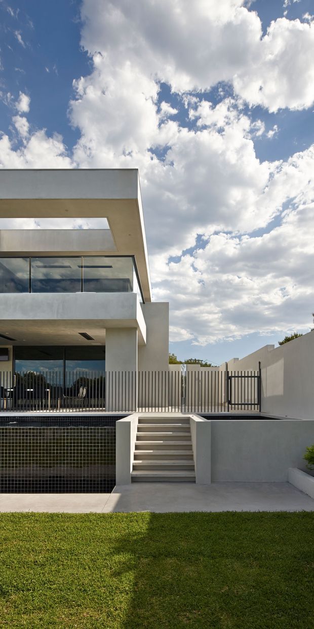 Exterior view of a contemporary multi-level residence with crisp white rendered walls, strong horizontal cantilevered roof forms, expansive glass openings, and dark vertical metal fencing. A tiled swimming pool occupies the foreground beside a concrete stairway and paved terrace, with a manicured lawn and bright cloud-filled sky.