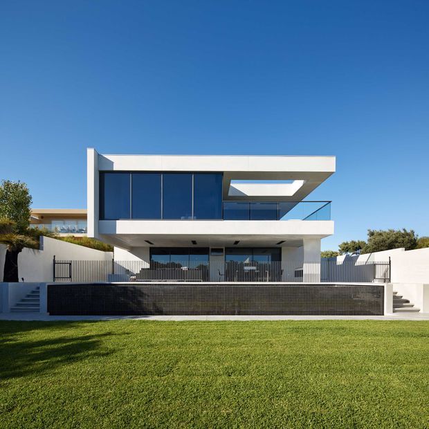 Front exterior view of a contemporary two-storey residence with crisp white rendered horizontal volumes, expansive dark glazing, a projecting upper-level terrace or balcony, and a prominent rooftop skylight or lightwell. The house sits behind a paved and fenced terrace with a dark tiled retaining wall, broad lawn, and steps at both sides. Clear blue sky and landscaped surroundings emphasize the clean, geometric facade.