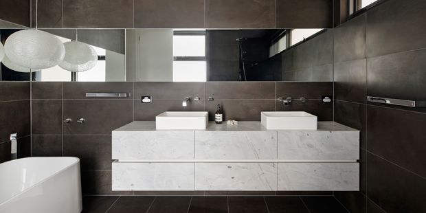 A contemporary bathroom with a double floating vanity, two white vessel basins, wall-mounted chrome tapware, large dark charcoal wall and floor tiles, and a broad white marble-look vanity surface. A freestanding white bathtub is partially visible at left, with a large horizontal mirror and sculptural pendant lights above the vanity.