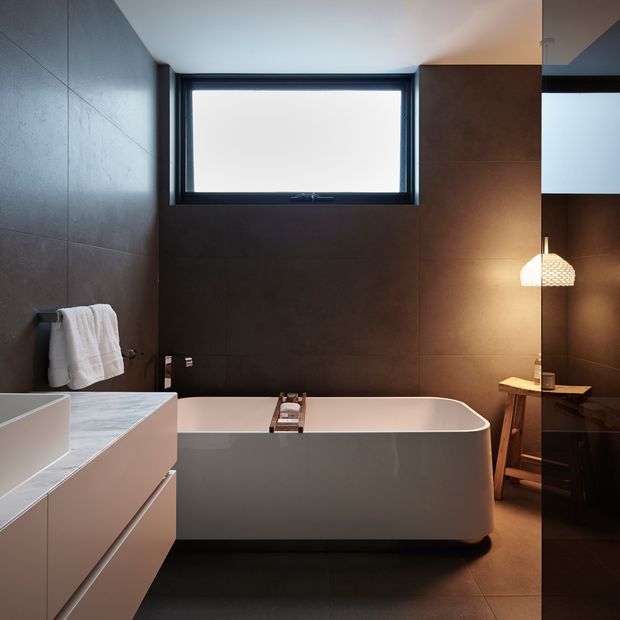 A dark, contemporary bathroom with a freestanding white bathtub, large dark brown wall tiles, a floating white vanity with pale stone benchtop, a black-framed horizontal window, towel rail, and warm pendant lighting reflected in a full-height mirror.