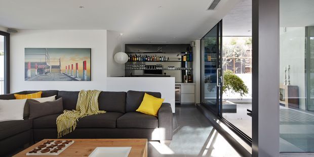 A bright, contemporary open-plan living room with a large charcoal sectional sofa, timber coffee table, artwork and yellow accent cushions. A compact built-in home bar with bottle and glass storage sits behind the seating area, while extensive black-framed glazing opens toward a planted courtyard or outdoor space. The room has a clean white interior, polished grey floor and strong natural light.