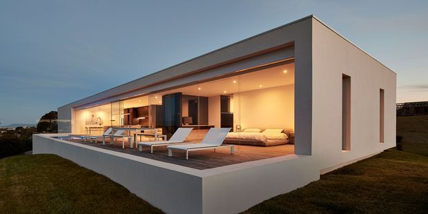 A contemporary single-storey flat-roof residence photographed at dusk, with a long open-plan glazed frontage opening onto a broad elevated terrace. The minimalist pale rendered facade has deep roof overhangs and narrow vertical side windows. Warm recessed lighting illuminates an interior living area and dining/kitchen zone, while the terrace includes outdoor seating, loungers and a low built-in seating area. The home sits in a grassy landscape with a distant coastal-looking horizon.