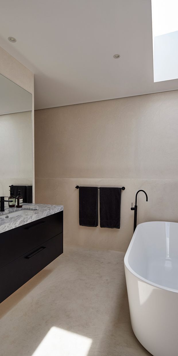 A bright, minimalist bathroom with a freestanding white bathtub, matte black wall-mounted tapware, black towels, and a dark floating vanity with a pale veined stone benchtop. Large mirror cabinetry is visible on the left, while a skylight and recessed ceiling lights provide natural and focused illumination. The walls and floor have a warm beige, softly textured finish.