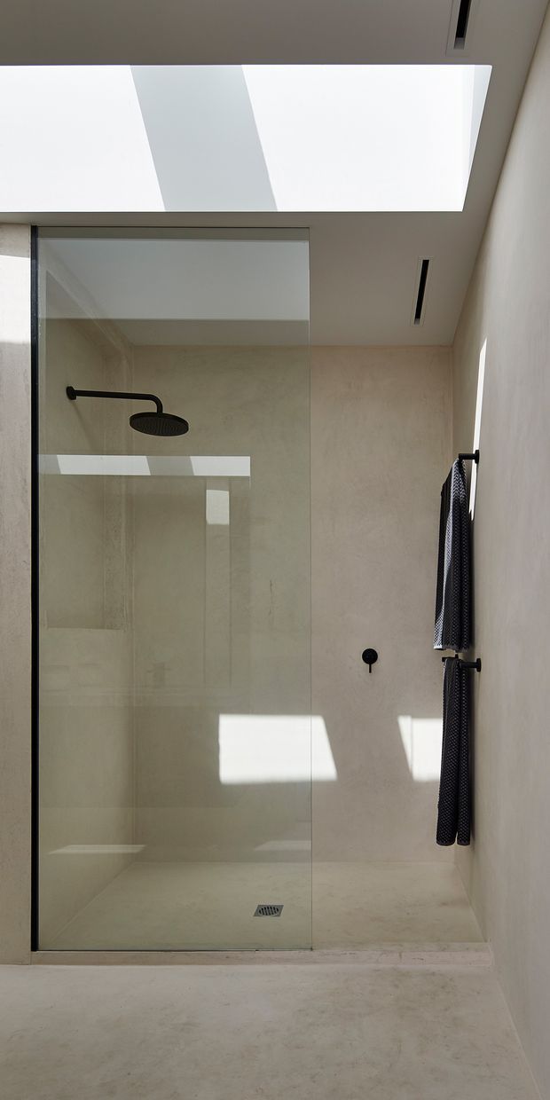 A minimalist walk-in shower bathroom with a frameless glass shower screen, large pale stone-look wall and floor surfaces, matte black shower hardware and towel rails, and a large skylight casting strong geometric sunlight into the space. The shower includes a rain showerhead, floor drain, and recessed ceiling ventilation or lighting slots.