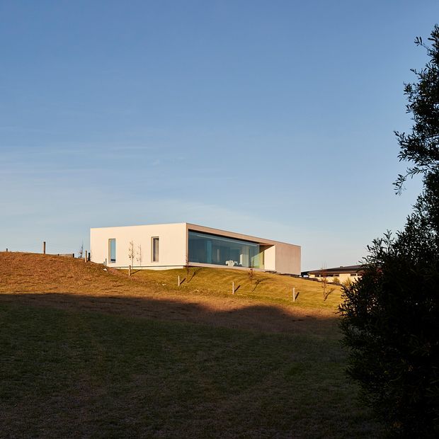 A low, minimalist contemporary house set on a gently sloping rural landscape. The building has crisp pale rendered walls, a mostly flat roofline, narrow vertical windows, and a large recessed glazed opening beneath a projecting roof or canopy. Dry grass, simple boundary fencing, and mature vegetation frame the house under a clear blue sky.