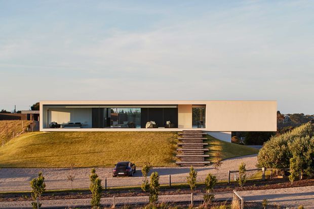 A low, minimalist contemporary house set on a landscaped sloping site. The long rectangular upper volume has smooth pale rendered walls, a flat roof, and extensive dark-framed floor-to-ceiling glazing opening onto a narrow terrace. A dramatic external stair of timber-toned treads descends across the grass to a gravel driveway, with structured planting in the foreground and rolling suburban landscape beyond.