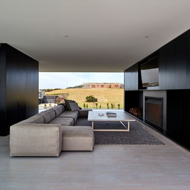 A modern living room with a large grey sectional sofa, low rectangular coffee table, dark built-in cabinetry, and an integrated fireplace. Floor-to-ceiling glazing opens the room to a broad landscaped rural view. The space has a restrained, monochromatic palette and clean linear detailing.