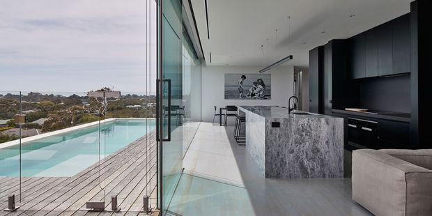 Contemporary open-plan kitchen and living area opening directly onto a poolside deck through extensive floor-to-ceiling glazing. The space features a large veined stone island, dark built-in cabinetry, pale flooring, streamlined recessed lighting, a dining area, and a partial sofa. The glazed facade provides broad suburban views and creates a bright indoor-outdoor connection.
