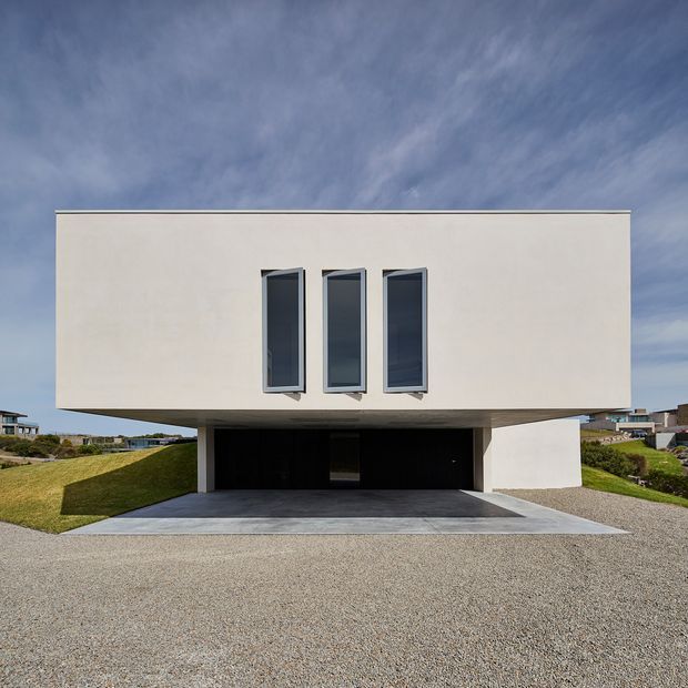 Front exterior view of a minimalist contemporary two-storey house with a large plain white rectangular upper volume projecting over a recessed double garage. Three tall narrow dark-framed windows punctuate the facade, while sloping lawns, gravel paving and neighboring homes surround the building.