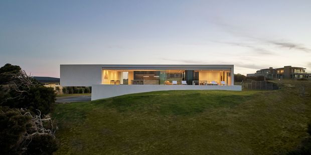Wide exterior view of a long, low-slung contemporary residence set into a gently sloping grassy site at dusk. The home has a crisp white rectangular facade, extensive warm-lit floor-to-ceiling glazing, a deep covered outdoor terrace, and a flat roof. Darker neighbouring residences and landscaped vegetation appear in the background.