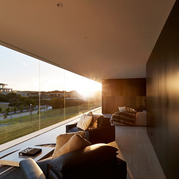 A warm, contemporary living room with floor-to-ceiling frameless glazing overlooking landscaped grounds. Dark leather seating, textured neutral cushions, timber wall panelling and a pale floor sit beneath a broad recessed ceiling with discreet downlights. Low evening sunlight creates strong golden illumination and reflections across the room.