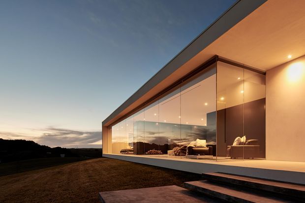 A dramatic dusk exterior view of a sleek modern single-storey residence. The building features a long glazed living area with floor-to-ceiling glass, warm recessed lighting, a projecting flat roof, and a minimal pale rendered facade with dark roof edging. Interior seating is visible through the glazing, while broad steps lead up from a paved path across a landscaped lawn.